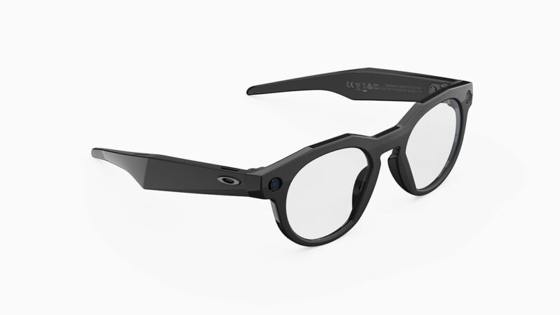 Black smart glasses with round clear lenses and thick arms, featuring a small button and the Oakley logo on the temple, displayed on a white background—now enhanced with Meta’s innovative technology.