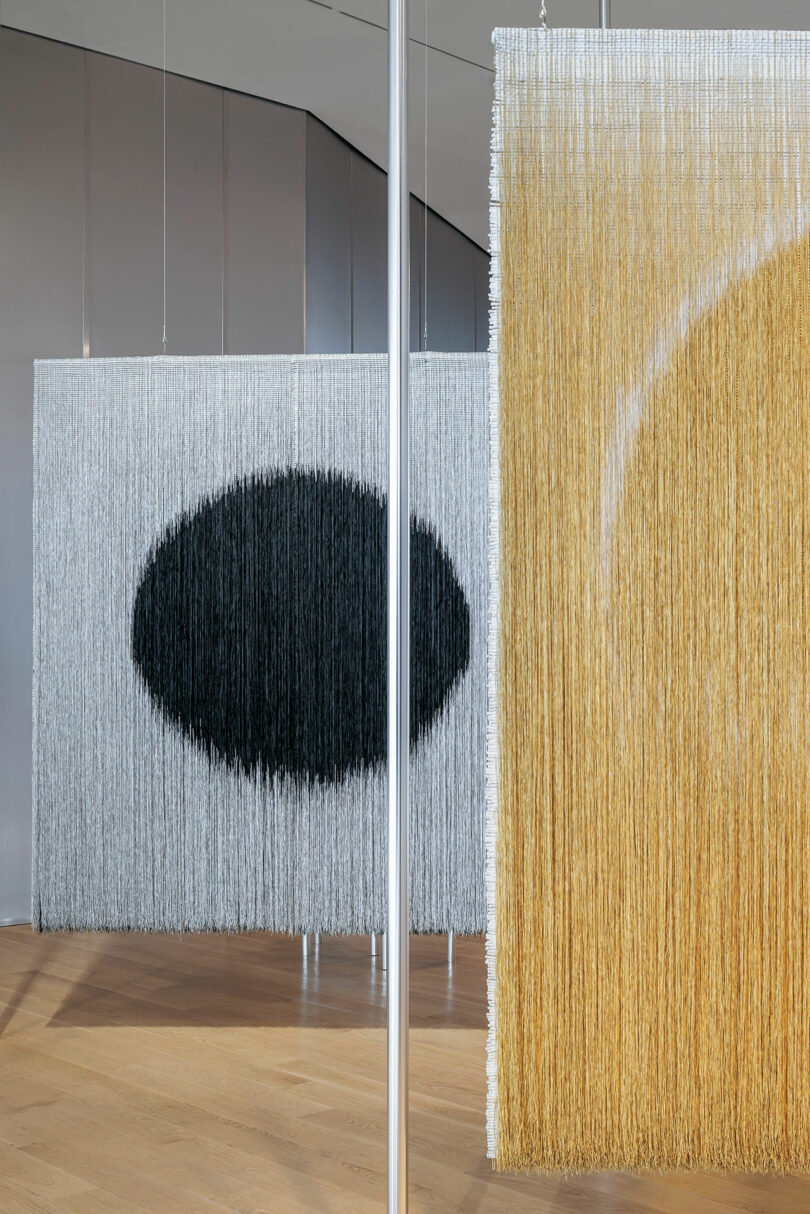 Hanging textile panels by Olga de Amaral made of vertical strings—one gray with a black oval, another yellow with a white arc—are displayed in a modern gallery space with wood floors.