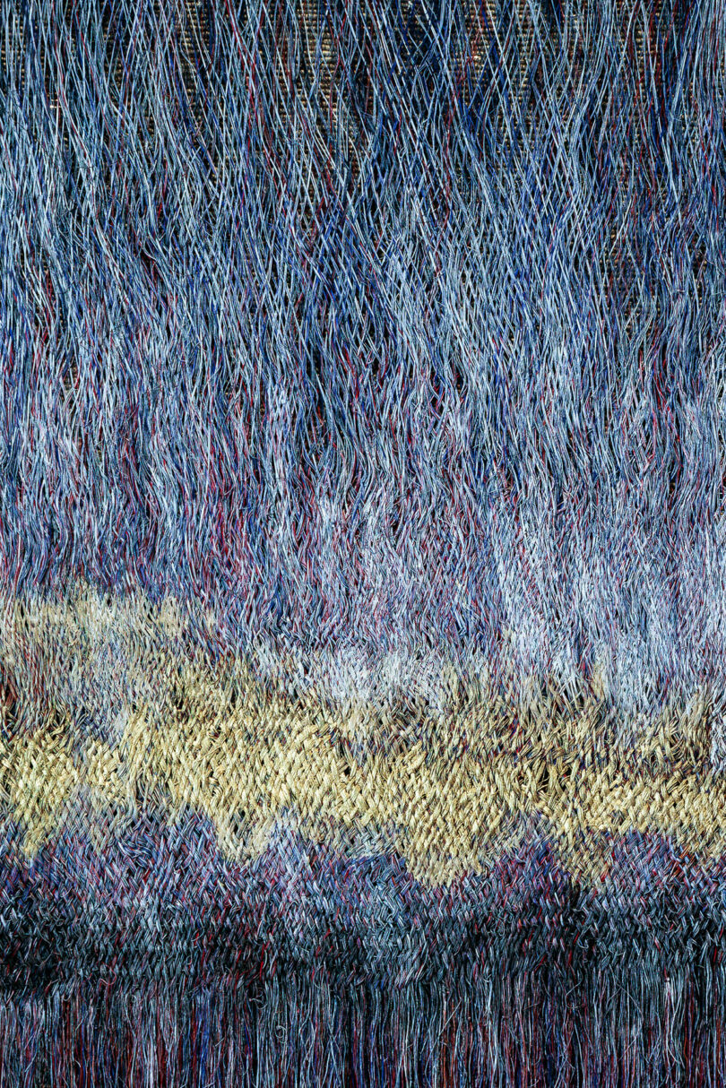 A close-up of a woven textile inspired by Olga de Amaral, featuring interlaced threads in blue, white, red, and tan that create a textured, wave-like pattern.