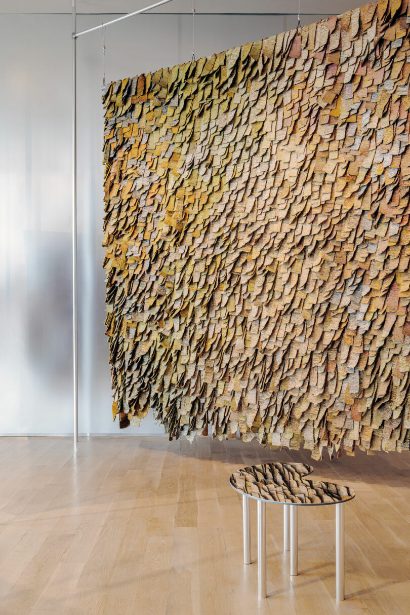 A hanging art installation inspired by Olga de Amaral, with layered, textured pieces resembling scales, floats above a small round table in a modern, minimalist room featuring sleek wood floors.