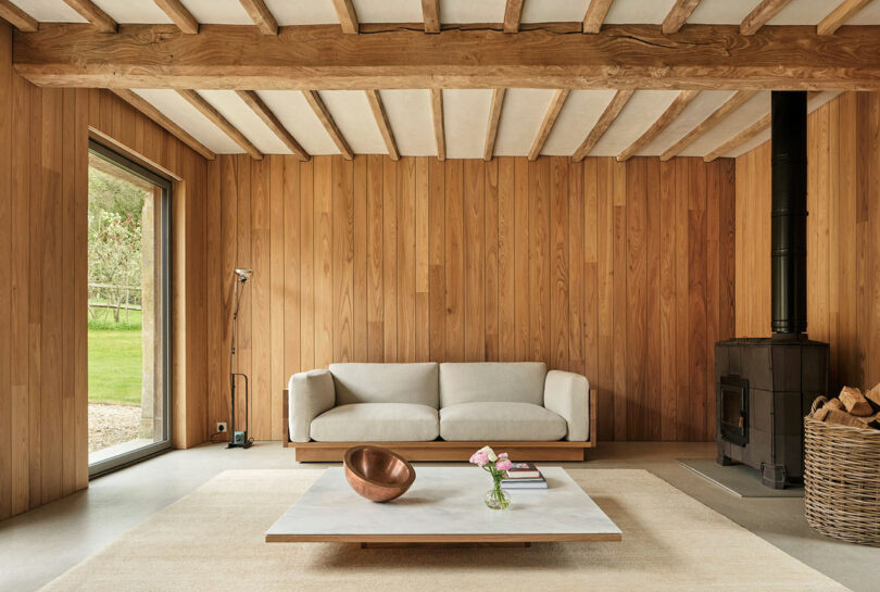 Minimalist living room with wooden walls and ceiling beams, a white sofa, a low coffee table with decor, a wood-burning stove, and a large window with a view of greenery