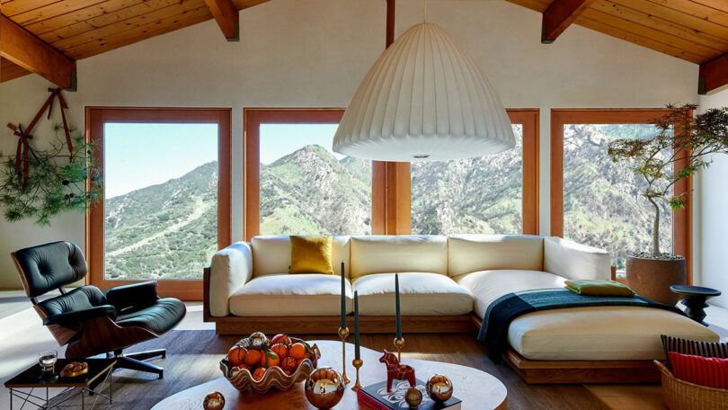 Modern living room with wooden ceiling beams, white sectional sofa, black lounge chair, large windows showing mountain views, and minimalist decor
