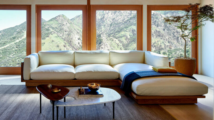 A modern living room with a cream sectional sofa, two coffee tables, a potted tree, and large windows overlooking green mountains