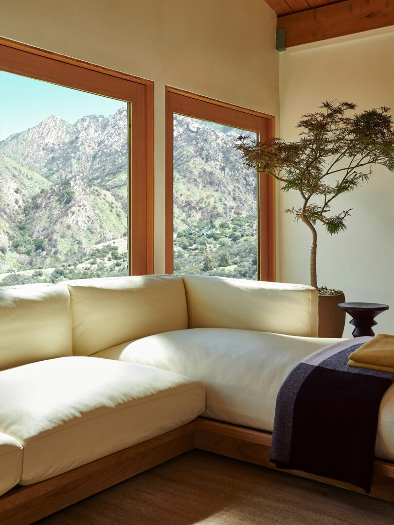 A modern living room with a cream sectional sofa, a potted plant, a side table, and large windows overlooking a mountain landscape