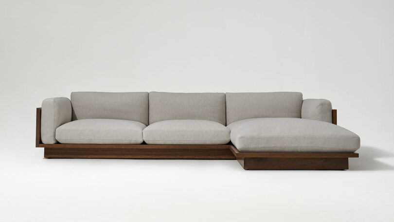 A modern sectional sofa with light gray cushions, a wooden base, and a chaise lounge on the right side, set against a plain white background