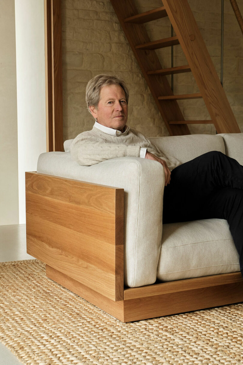 A man with gray hair, wearing a light sweater and dark pants, sits on a beige sofa with wooden panels in a modern room with wooden stairs and a woven rug