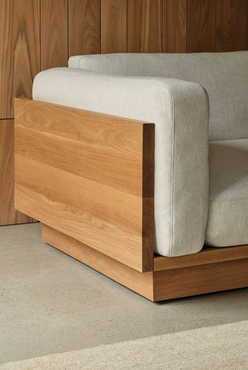 A modern armchair with light gray upholstery and wooden side and base panels, set against a wooden wall and placed on a light stone floor