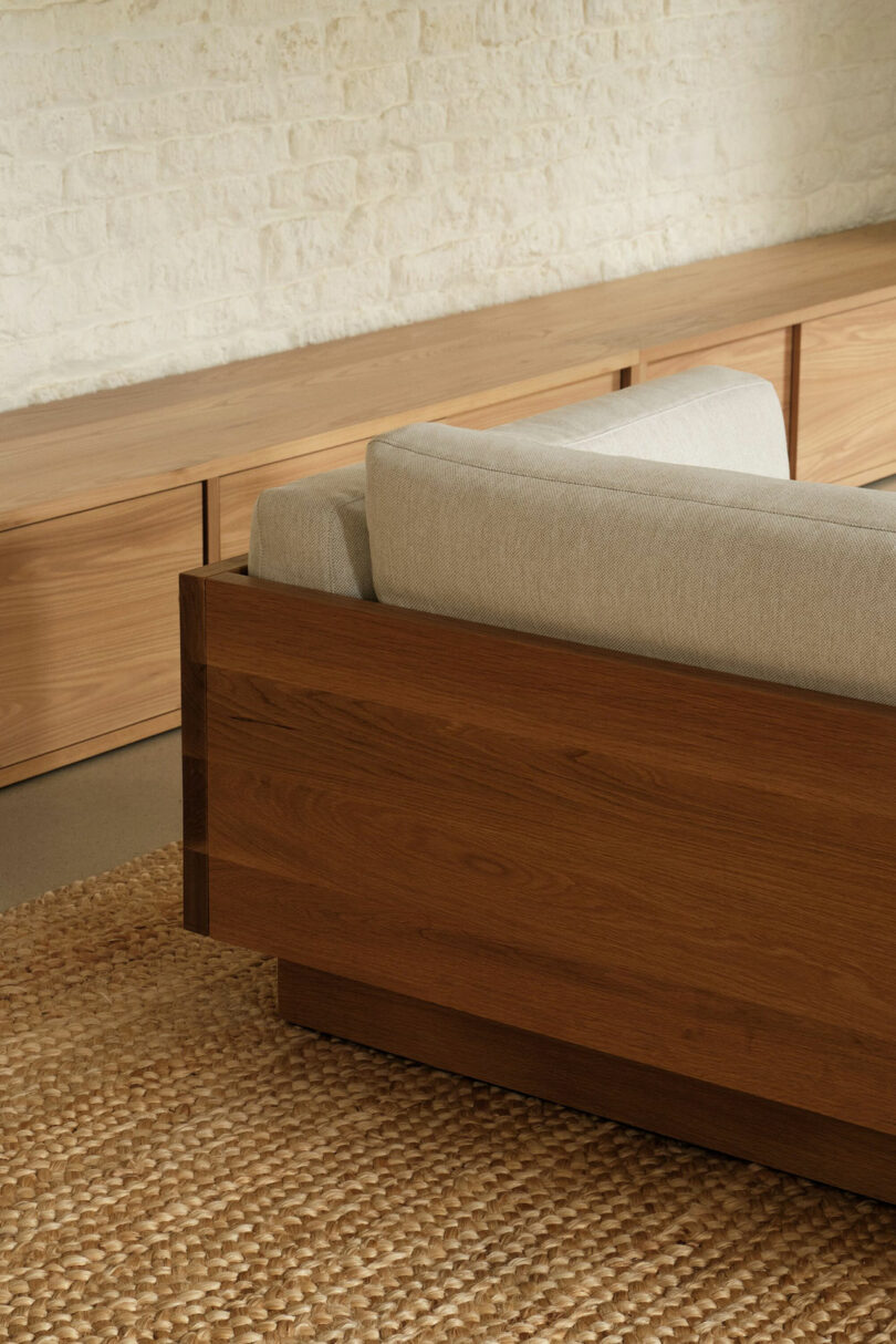 A wooden sofa with light beige cushions is placed on a woven rug beside a long, low wooden cabinet against a white brick wall