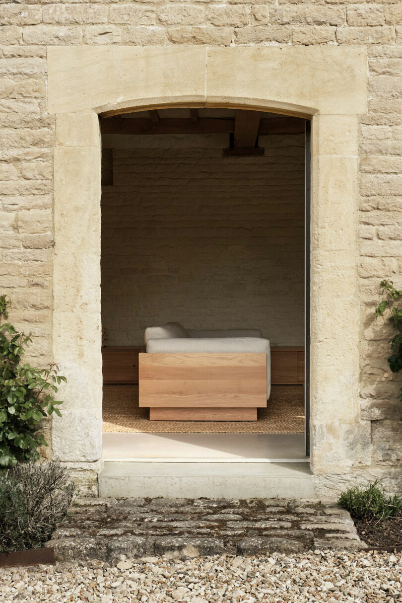 A wooden-framed sofa sits in a minimalist room with stone walls, viewed through an arched doorway of a rustic stone building