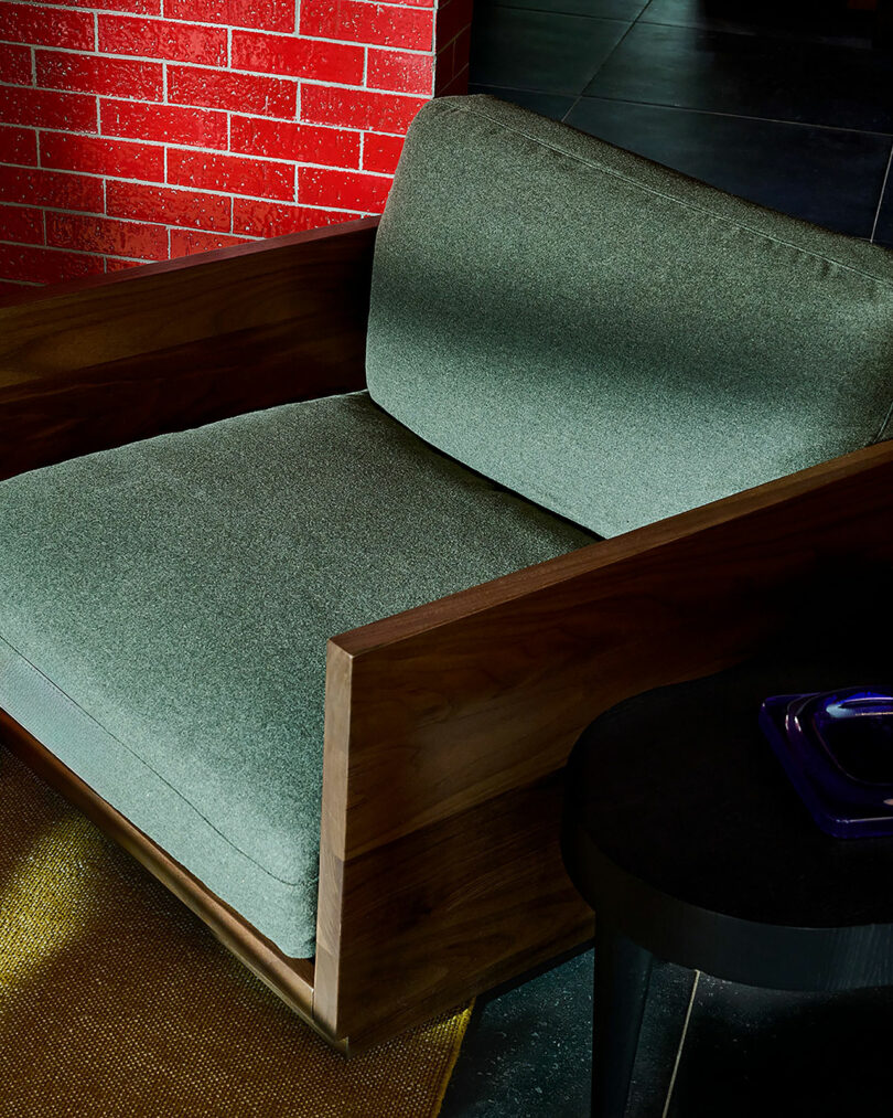 A modern armchair with green upholstery and wooden frame next to a round black side table, set against a red brick wall and dark tile flooring