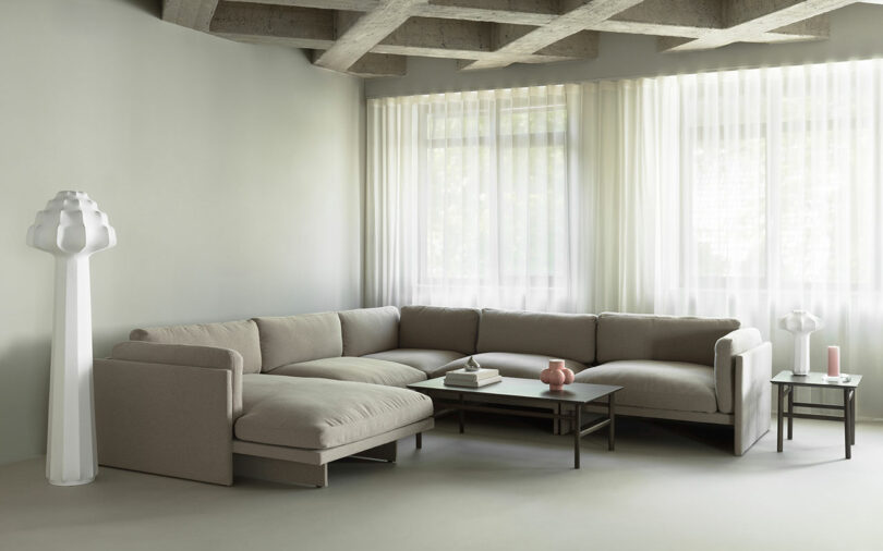 A modern living room with a beige sectional sofa, two coffee tables, a white sculptural floor lamp, a small table lamp, and sheer curtains over large windows.