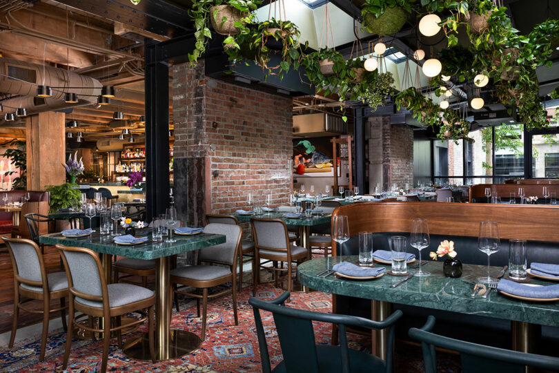 Modern restaurant interior with green marble tables, wooden chairs, exposed brick walls, hanging plants, and decorative lighting. Tables are set with glassware and napkins.