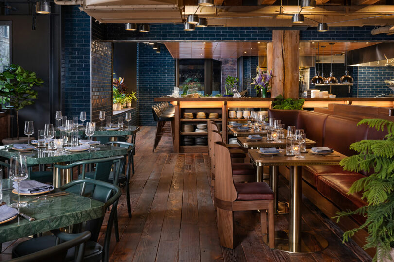 Modern restaurant interior with wooden floors, green marble tables, leather booths, set tables with glassware, and an open kitchen in the background.