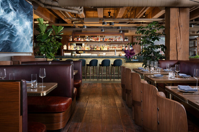 Modern restaurant interior with wooden floors, leather booths, and tables set with glassware. A bar with high stools is in the background, surrounded by plants and shelves of bottles.