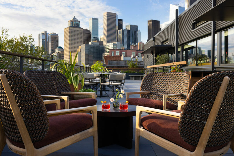 A rooftop patio with four wicker chairs around a small table, city skyscrapers in the background, and drinks set on the table.