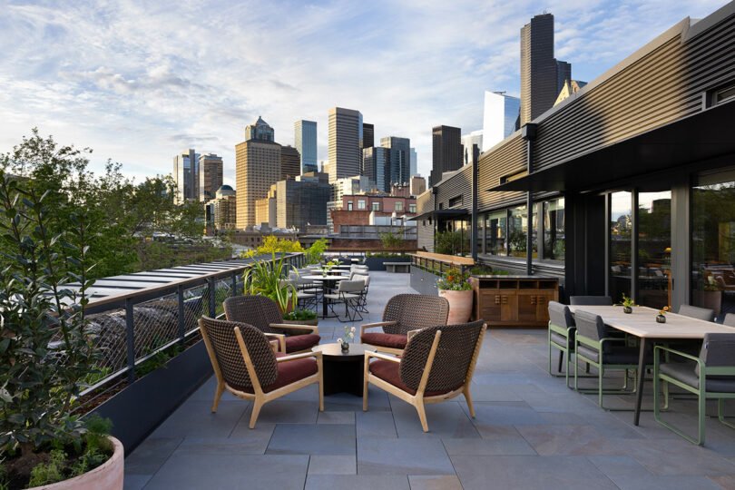 A modern rooftop patio with outdoor seating overlooks a city skyline with tall buildings under a partly cloudy sky.