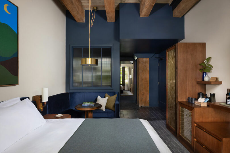 Modern hotel room with a bed, blue accent wall, wooden ceiling beams, a seating area with table, and a cabinet with drinks and decor. A landscape painting hangs above the bed.