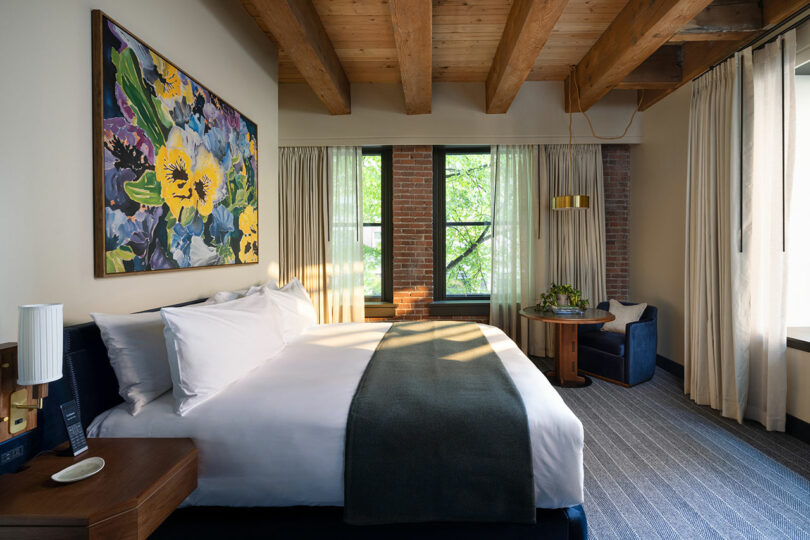 A modern hotel room with a king bed, large floral artwork above the headboard, exposed wooden ceiling beams, tall windows, and a small seating area with a chair and table.