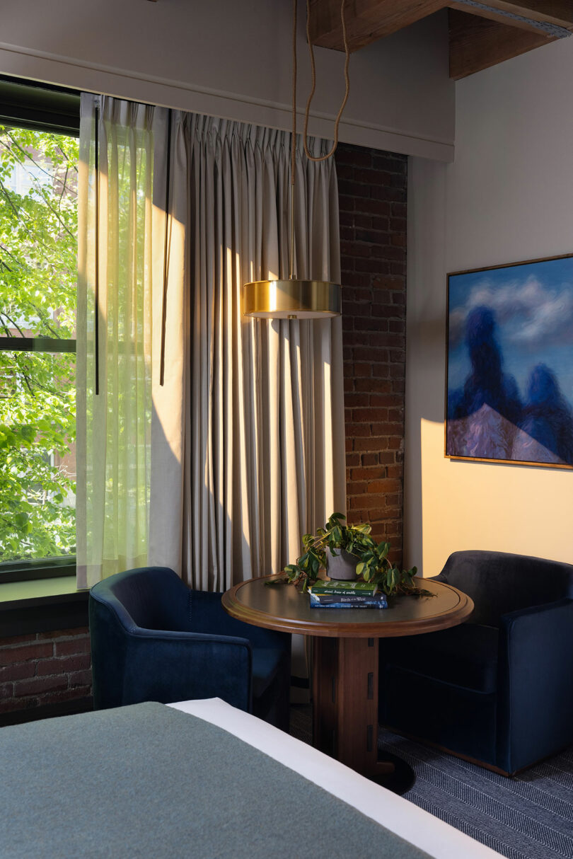 A small round table with two blue chairs sits by a window with sheer curtains. A potted plant and books are on the table, and a painting hangs on an exposed brick wall.