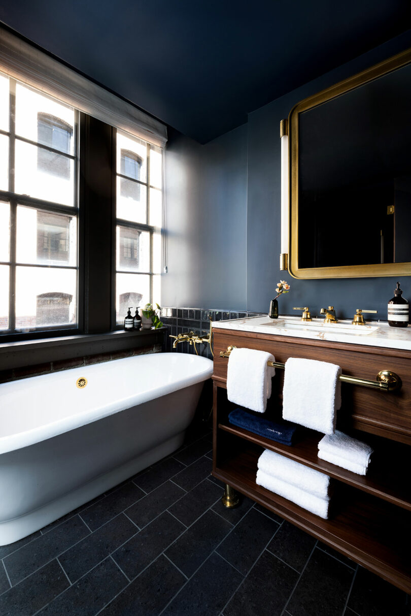 Modern bathroom with a freestanding tub, dark tile floor, wooden vanity with towels, and large mirror; natural light streams in through tall windows.