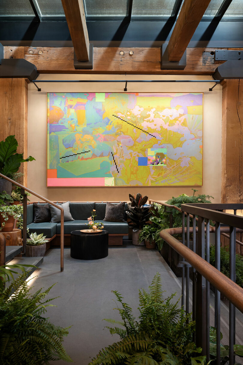 Modern indoor lounge area with a large abstract painting on the wall, surrounded by greenery, wooden beams, and contemporary seating.