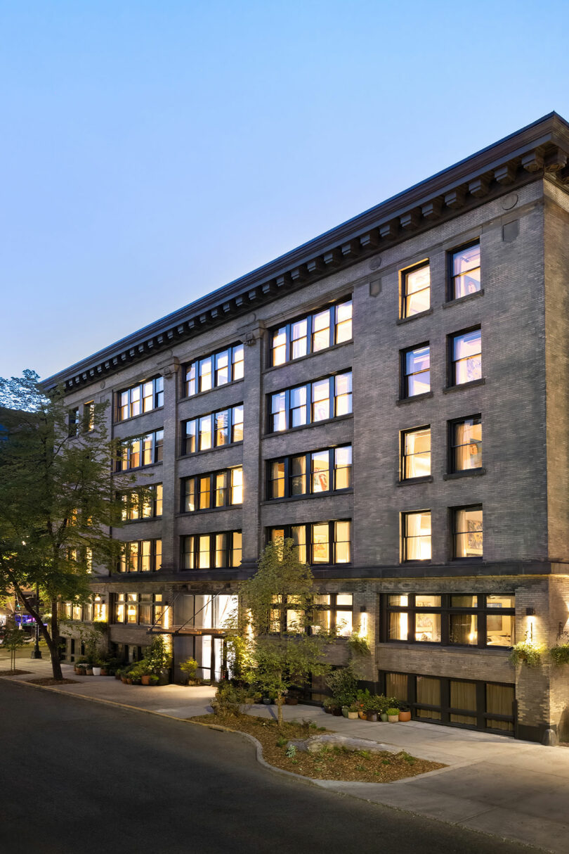 A five-story brick apartment building with large windows illuminated from inside, set on a tree-lined street at dusk.