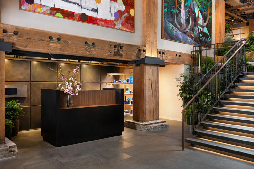 Modern lobby with a black reception desk, wooden beams, metal staircase, indoor plants, and colorful abstract artwork on the walls.