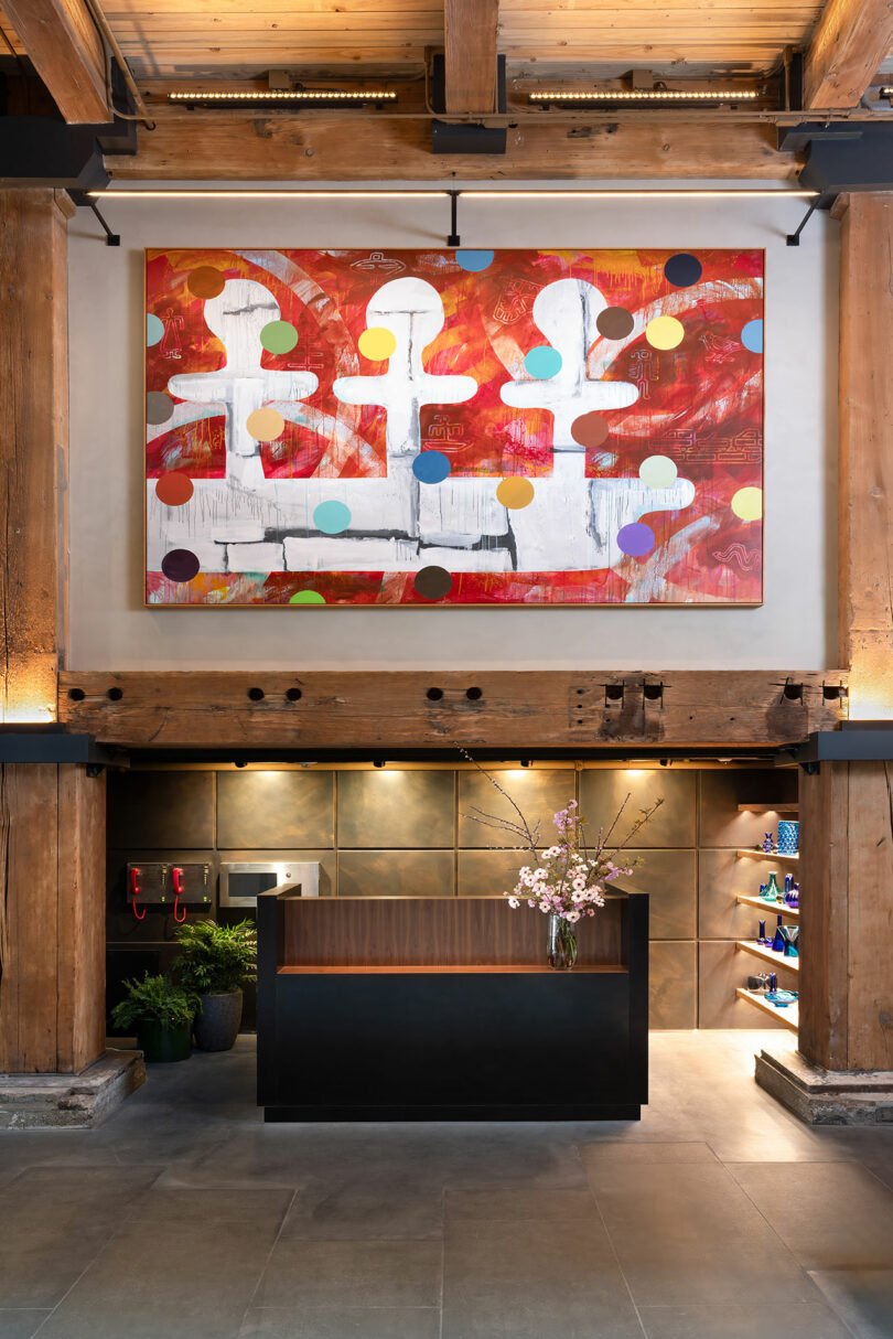 A modern reception area with a small dark desk, flower arrangement, wooden beams, wall shelves, and a large abstract painting with red, white, and colorful dot patterns above.