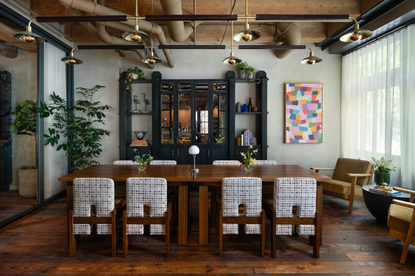 A modern dining room with a wooden table, eight patterned chairs, a black cabinet, large plants, abstract wall art, and exposed ceiling pipes. Natural light enters through sheer curtains.