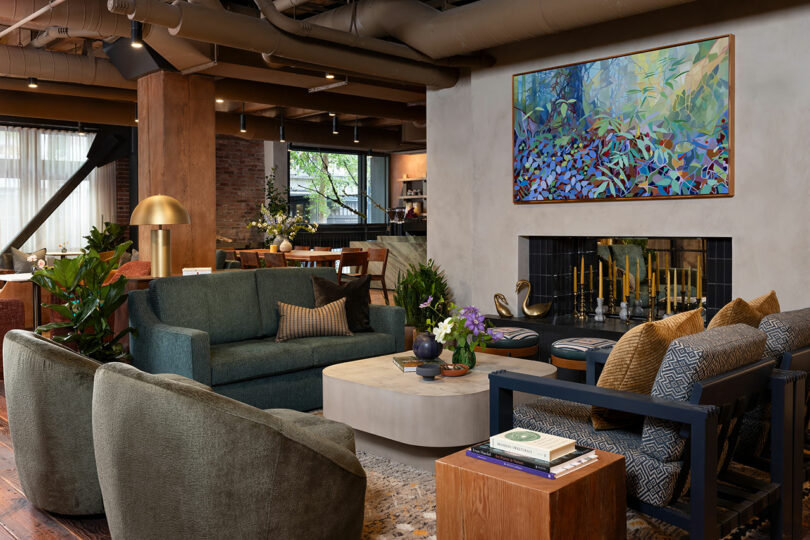 Modern living room with green and blue seating, a coffee table, plants, and a fireplace beneath a colorful abstract painting. Exposed ceiling pipes and large windows are visible in the background.