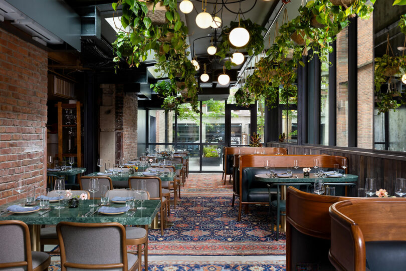 A modern restaurant interior with green marble tables, wooden booths, patterned rug, hanging globe lights, and ceiling plants, featuring large windows and an exposed brick wall.