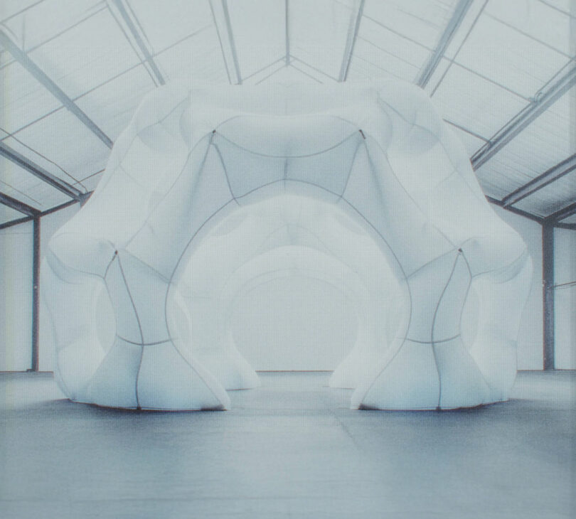 A large, white, futuristic structure with curved arches stands inside a spacious warehouse with a high, glass-paneled ceiling.