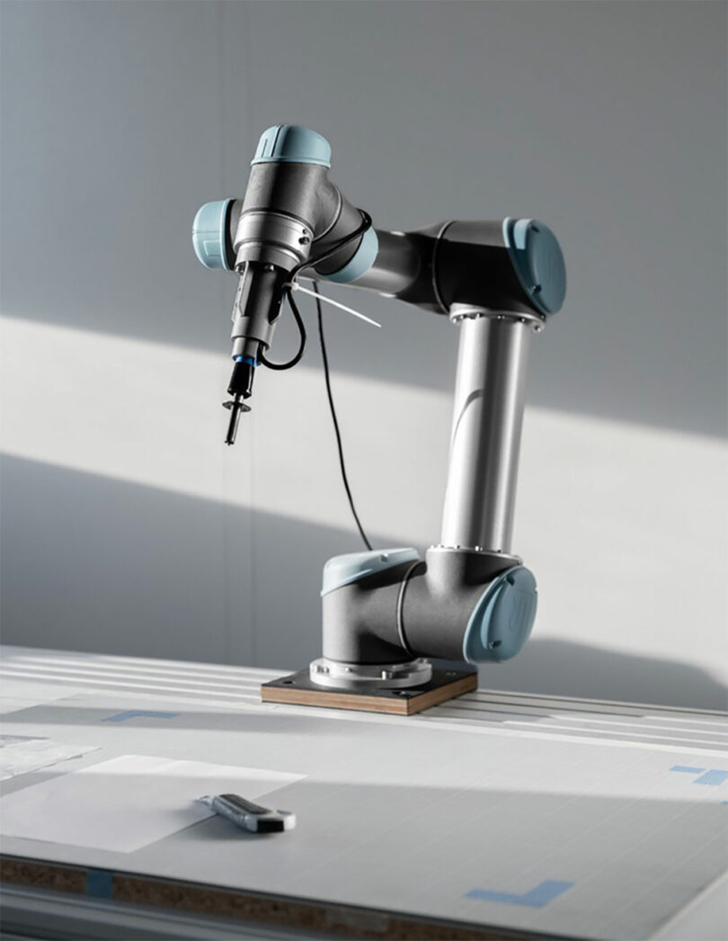 A robotic arm with a tool attached is positioned on a work table, with papers and a pen nearby under natural light.