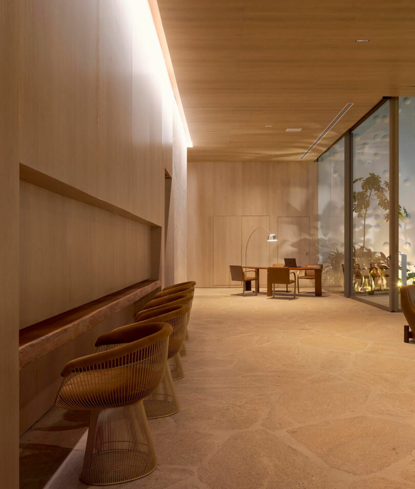 Spacious interior with light wood walls, stone floor, modern wire chairs along one wall, and a desk with a lamp near large floor-to-ceiling windows.