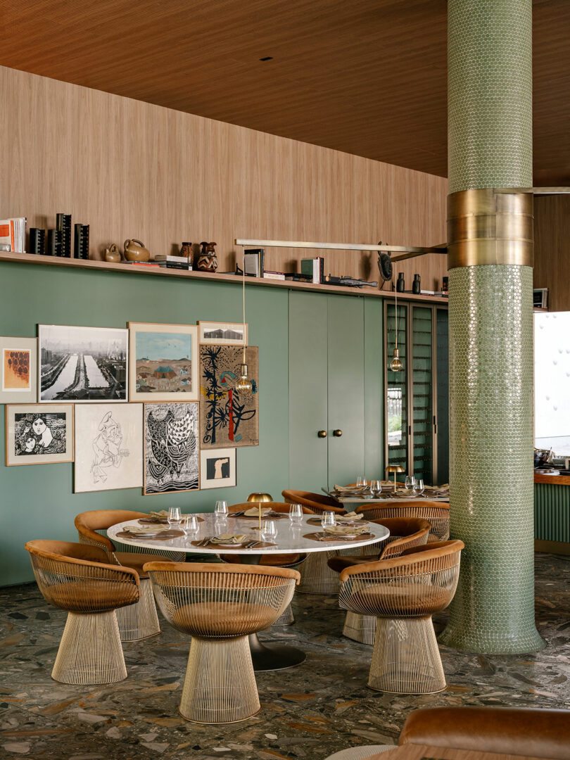 A stylish restaurant interior with a round marble table, wicker chairs, green paneled walls, eclectic art, a mosaic column, and decorative objects on a high shelf.