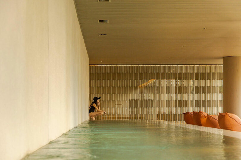 A person in a swimsuit and hat sits at the edge of an indoor pool with calm water, beside beige walls and lounge chairs.