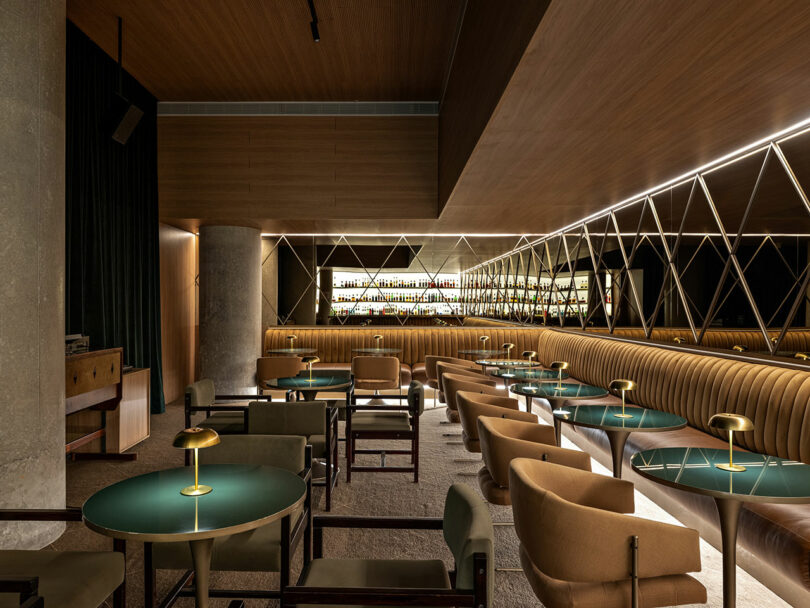 A modern bar interior with green tables, tan upholstered chairs, a long banquette, mirrored wall panels, and backlit shelves displaying bottles.