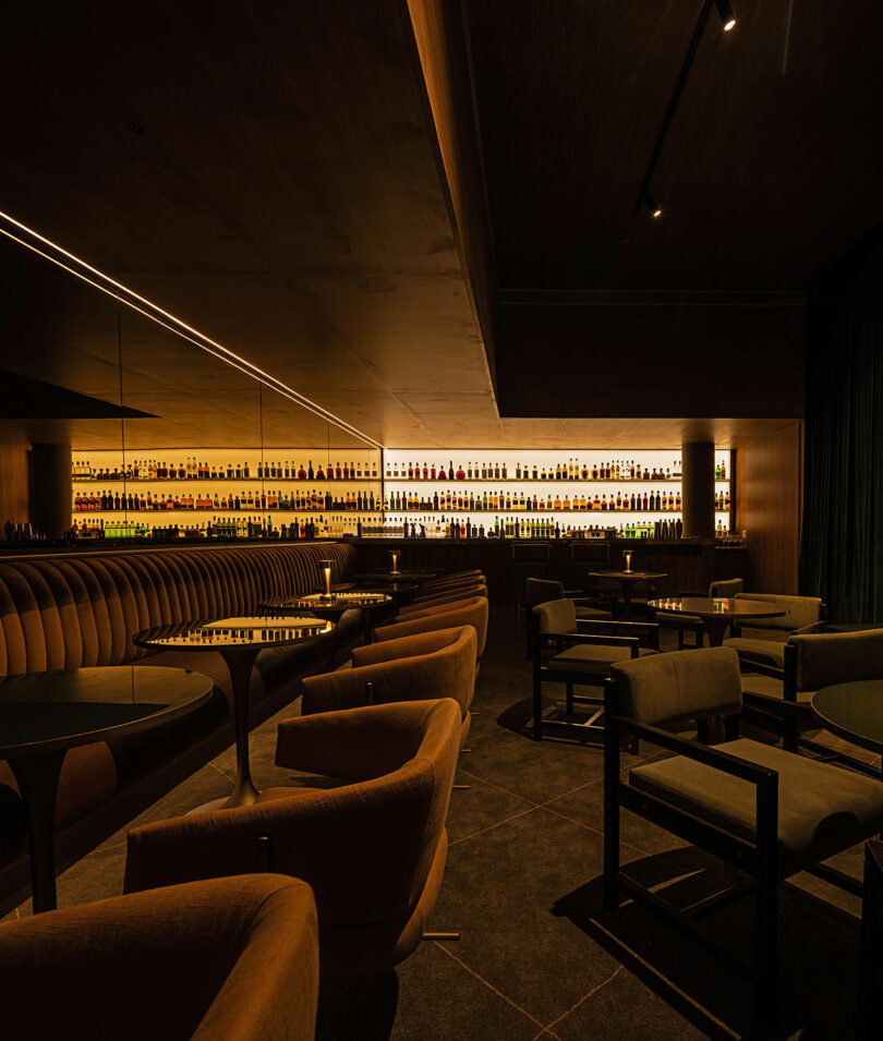 A dimly lit, modern bar with curved brown chairs, round tables, and shelves of backlit liquor bottles against the wall.