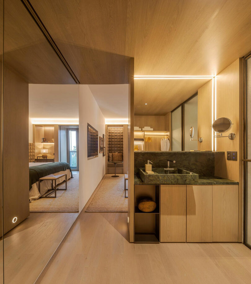 Modern hotel suite with a view of a bedroom and an open bathroom area featuring a marble sink, wooden surfaces, and warm ambient lighting.