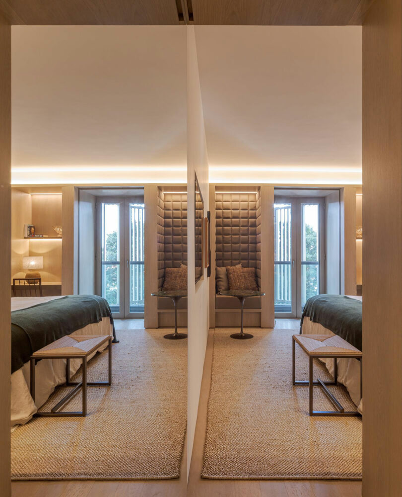 A modern bedroom with a central dividing wall, creating a mirrored effect with two beds, benches, and cushioned seating areas by large windows.