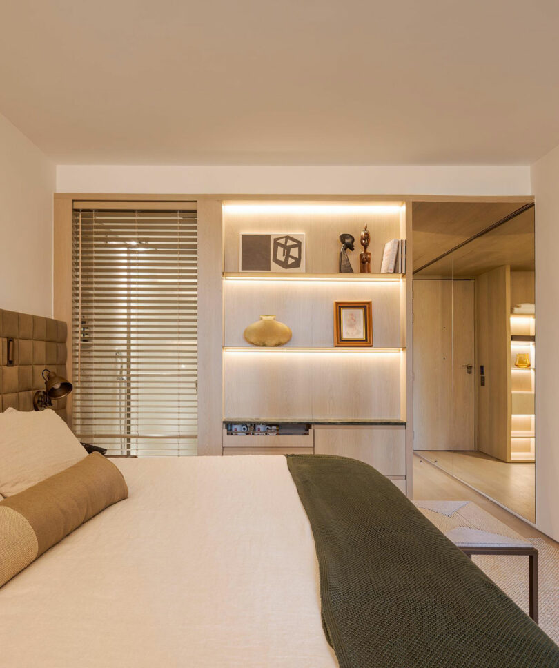 A modern bedroom with a beige bed, green blanket, built-in shelves with decor items, wooden blinds, and a mirrored wall reflecting a hallway.