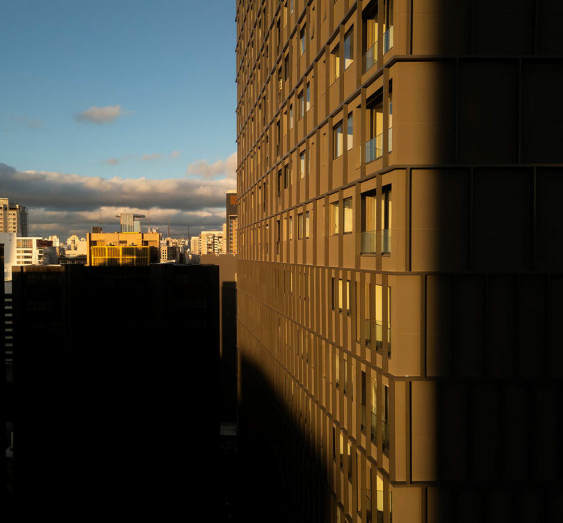 A tall building partially in shadow with sunlight illuminating its side, against a cityscape background under a cloudy sky.