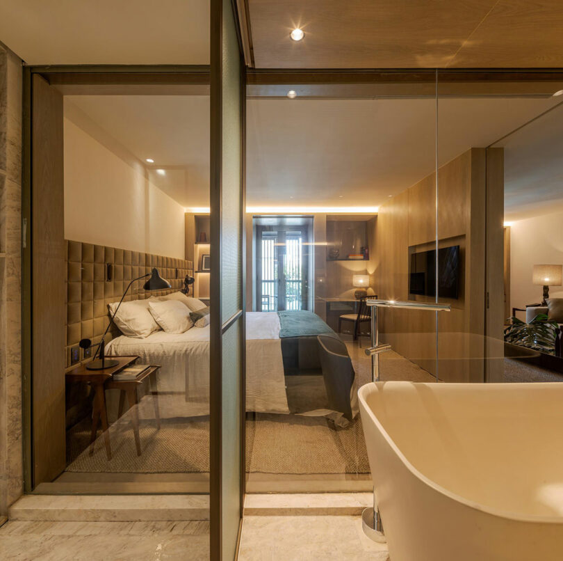 A modern hotel room with a glass partition separating a freestanding bathtub from a bedroom featuring a bed, bedside tables, and a TV on a wooden accent wall.