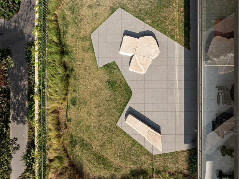 Aerial view of a geometric patio with stone benches and tables, surrounded by grass and plants, adjacent to a glass building and a walkway.