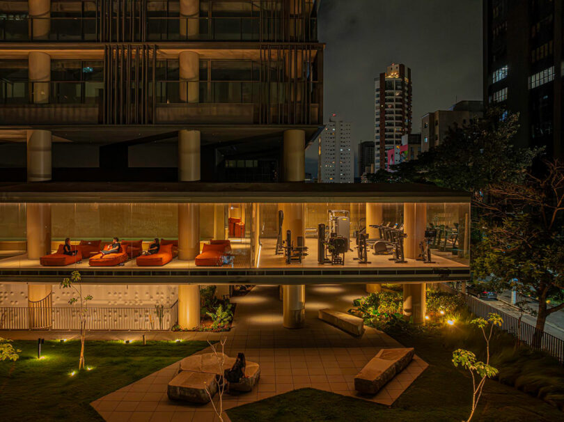 A modern building at night with a well-lit gym and lounge area on an upper floor, visible through large windows; landscaped grounds with seating are in the foreground.