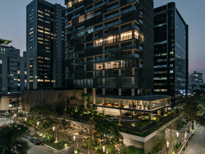 Modern high-rise building with illuminated windows and a landscaped area at its base, surrounded by other tall buildings at dusk.