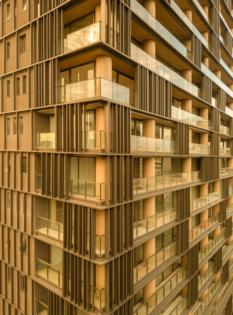 Modern building facade with numerous glass balconies, vertical and horizontal metal beams, and beige columns reflecting warm sunlight.