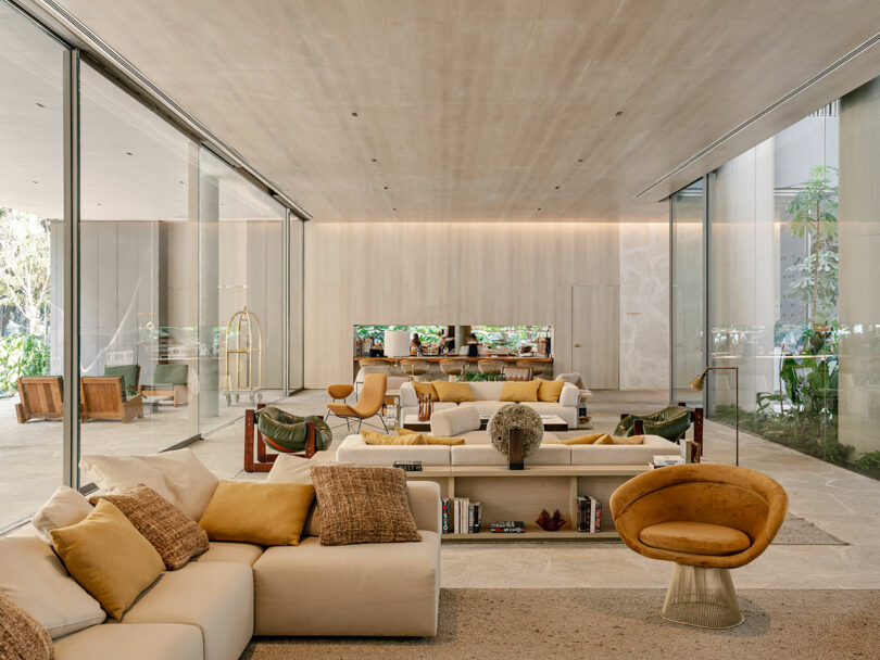Spacious modern living room with beige and yellow furniture, large glass walls, natural light, and garden views. Books and decorative items are on a central white shelving unit.