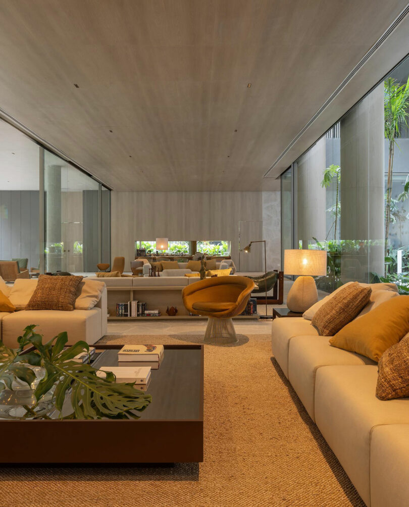 Modern living room with neutral-toned sofas, yellow accent pillows, a glass coffee table, large windows, and indoor plants, creating a bright and spacious atmosphere.