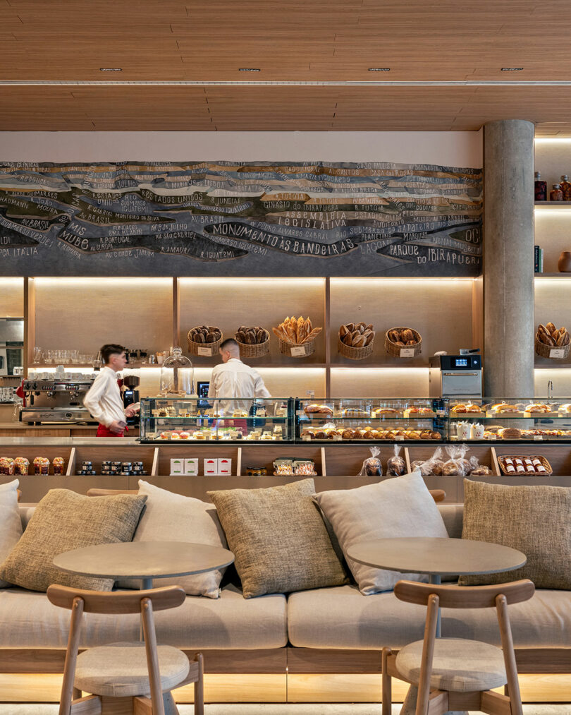 Modern bakery café interior with two staff behind a glass display of pastries, shelves of bread, and cushioned seating in the foreground.